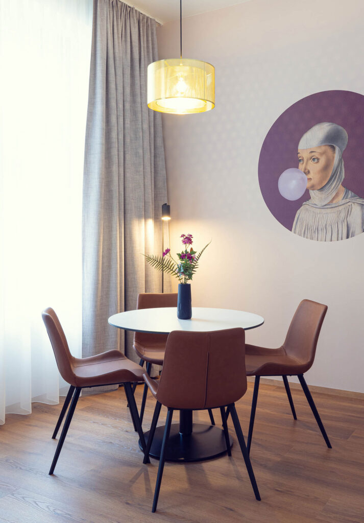 Runder, weißer Tisch mit drei braunen Stühlen. Es leuchtet eine Deckenlampe